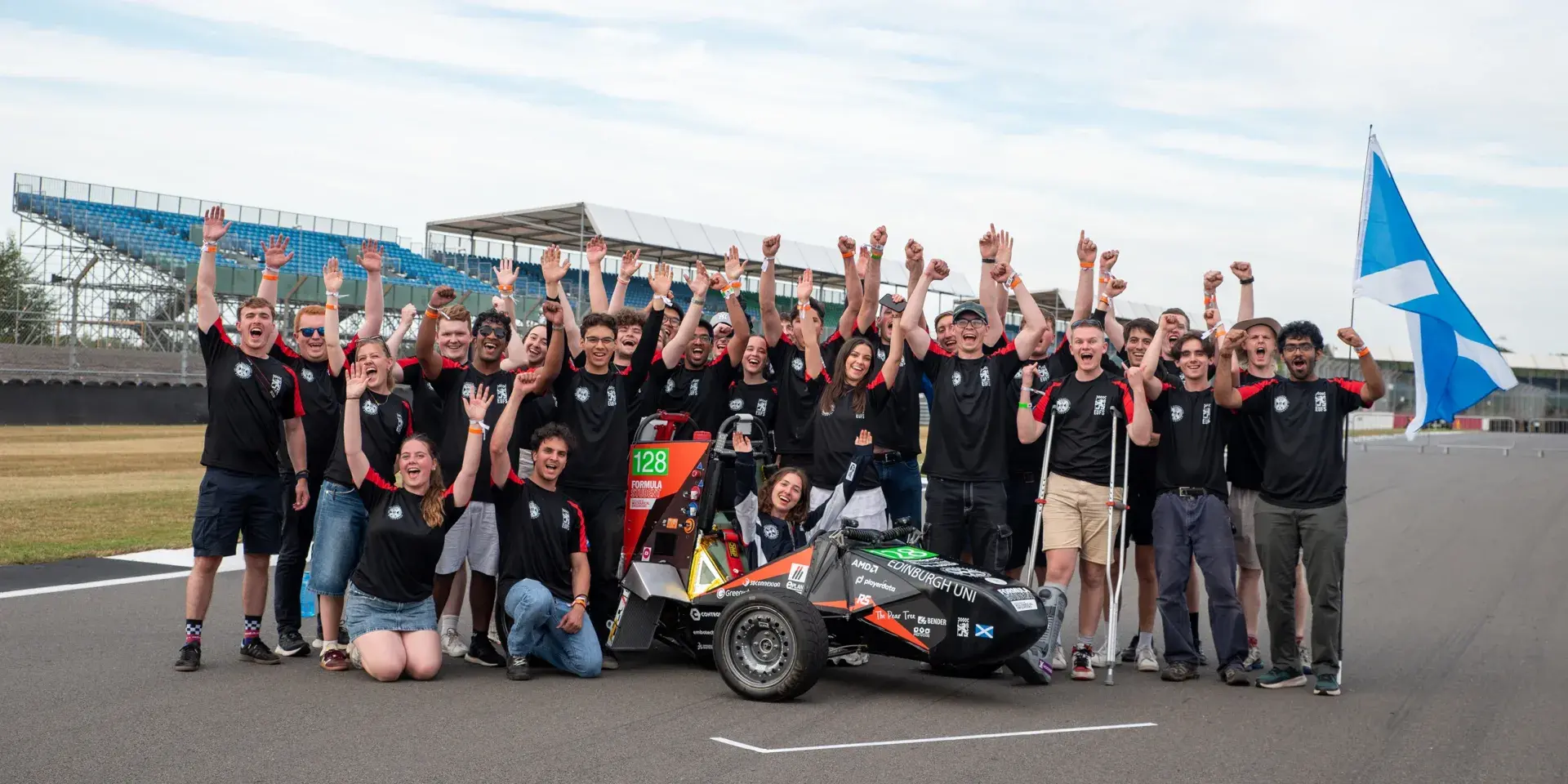 Group of people on a racetrack holding their hands up happily with a formula car in the middle and a Scottish flag on the right