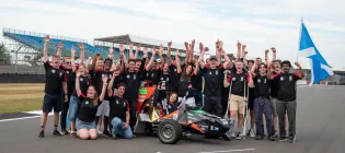 Group of people on a racetrack holding their hands up happily with a formula car in the middle and a Scottish flag on the right